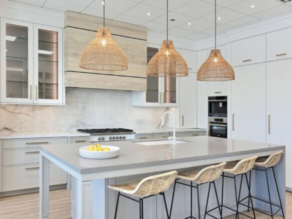 Modern kitchen with a large gray island, featuring three rattan pendant lights and four wicker stools. White cabinets and a marble backsplash create a stunning backdrop. On the island countertop sits a bowl of bananas, perfect for those exploring new homes or pre-construction options in the GTA.