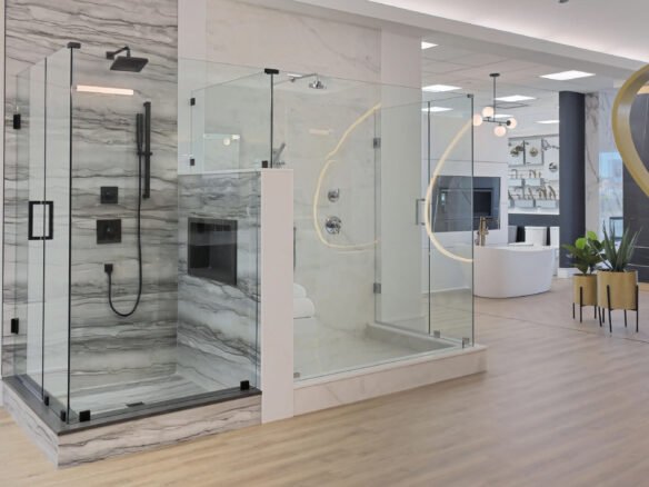 Discover a modern bathroom showroom, perfect for new homes, featuring a glass-enclosed shower with gray and white marble walls and black fixtures. Adjacent is a curved glass section. In the background, contemporary fixtures accompany a white bathtub on elegant wooden flooring.