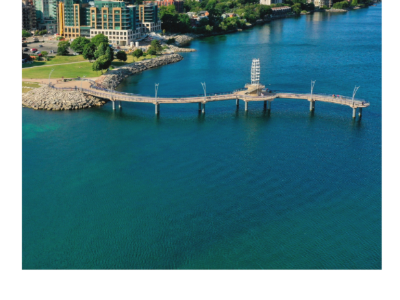 An aerial view of a coastal city with a long pier extending into a blue lake. The foreground showcases the pier and surrounding water, while residential and commercial buildings line the shore. New homes dot the skyline under a sky adorned with clouds.