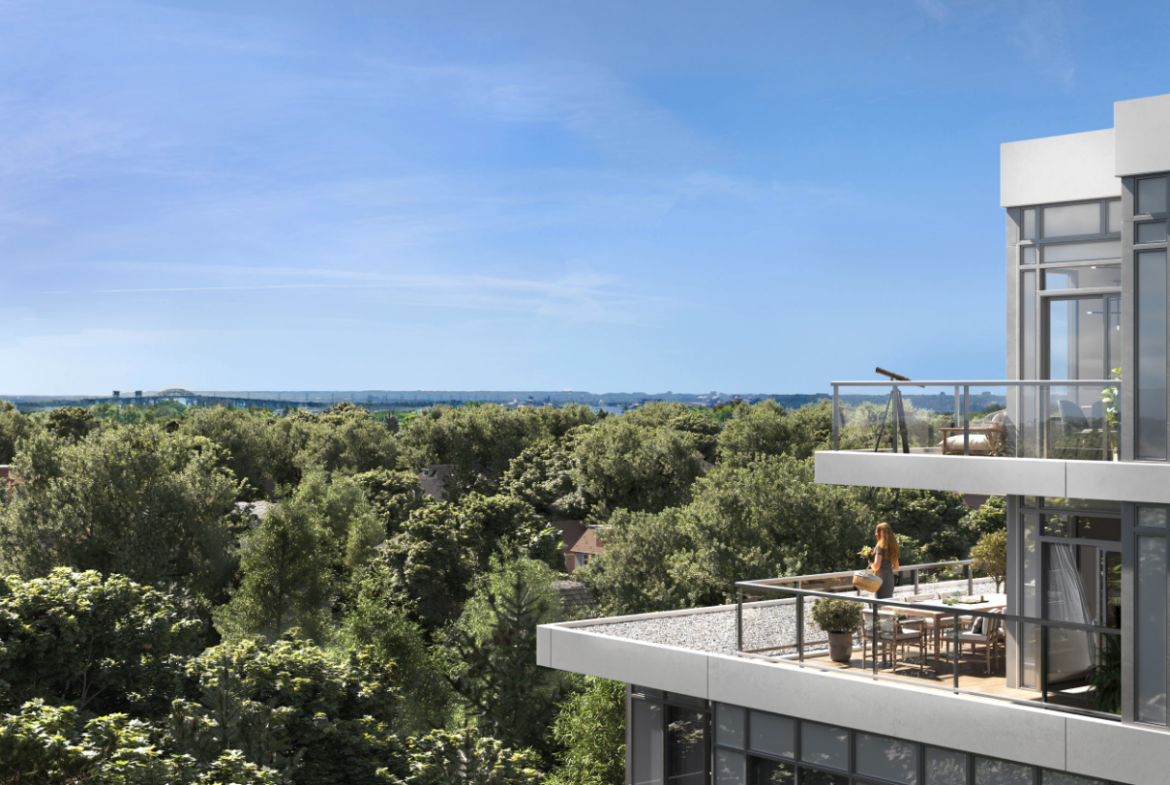 A modern, multi-story building with large glass windows and balconies overlooks a lush green forest. A person stands on a balcony enjoying the view under a clear blue sky, epitomizing the appeal of new homes in Brampton for those craving a scenic escape.