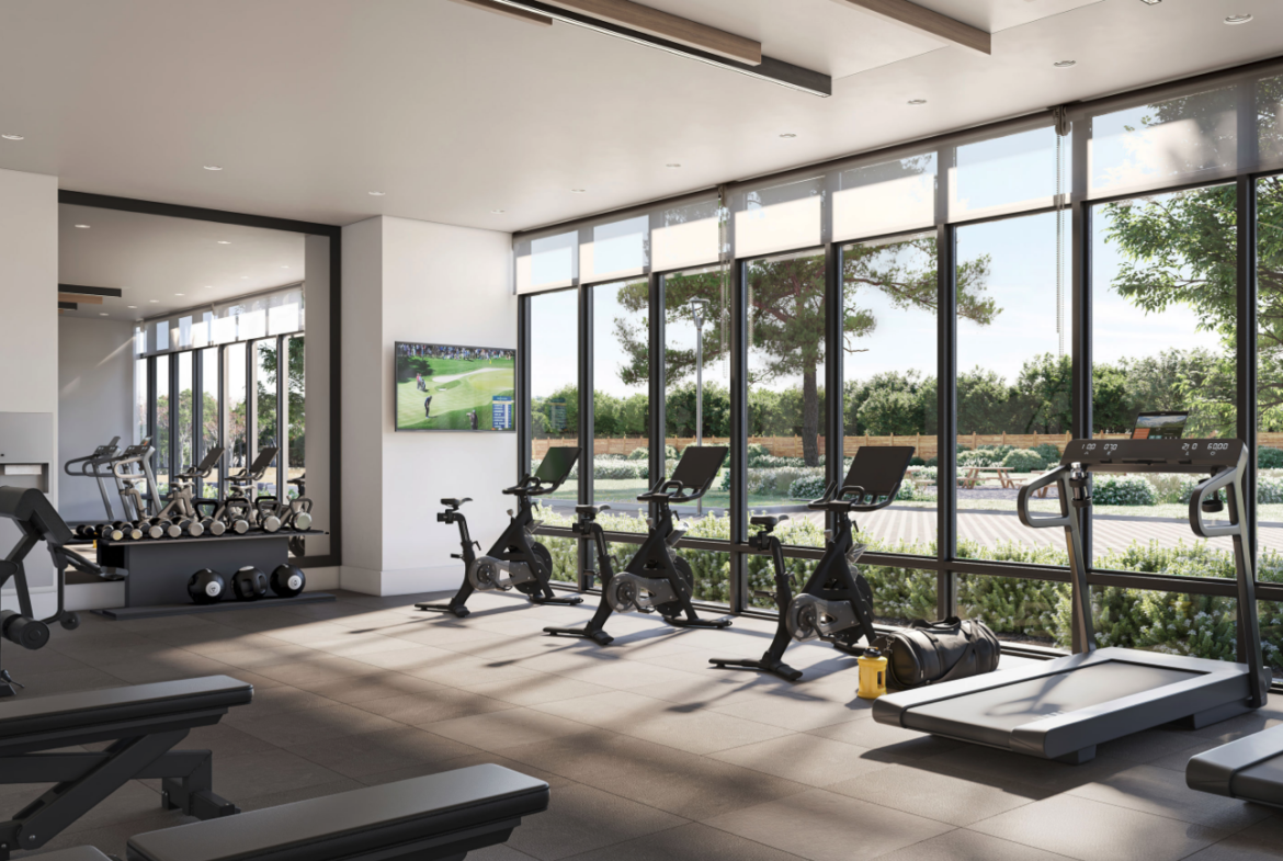 A modern gym with large windows offers a leafy view and includes exercise bikes, treadmills, and weightlifting equipment. Perfect for residents of new homes in Toronto, a TV displays sports while natural light fills the room, highlighting a clean and organized space.