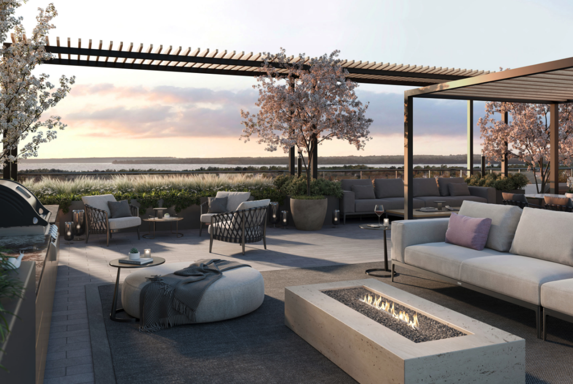 A modern rooftop patio in one of the new homes in Toronto features comfortable seating around a rectangular fire pit. Blossoming trees and potted plants decorate the space. The setting sun casts a warm glow over the relaxed ambiance, with a view of nature in the background.