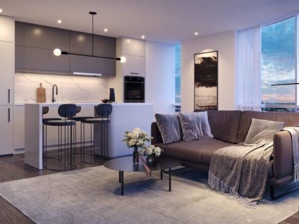 A modern living room at Park Place VMC boasts a sleek kitchen. The space features a brown sofa with gray pillows, a coffee table with flowers, two bar stools at the island, and large windows showcasing a sunset view. The decor is minimalist and elegant.