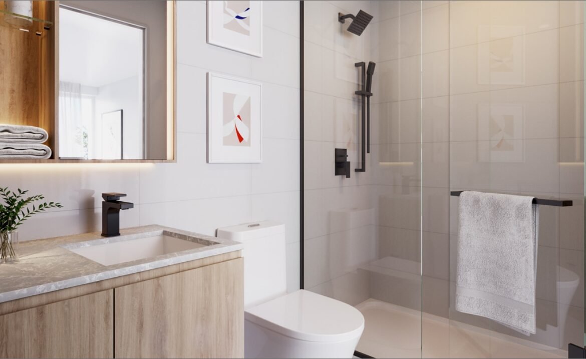 This modern bathroom in the new condos in GTA features a wooden vanity, white countertop, and sleek black fixtures. A large mirror and framed art adorn the wall, while a glass shower door holds a crisp white towel. Natural light floods through a window, enhancing the clean and minimalist design.