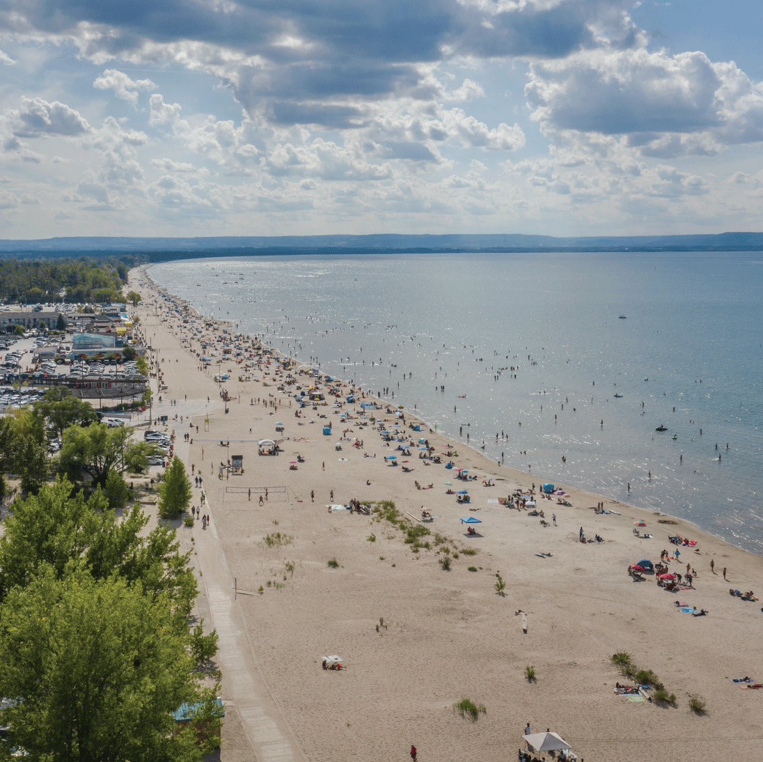 Aerial view of a large, sandy beach with people relaxing and swimming. The beach is dotted with umbrellas and towels under a partly cloudy sky. The ocean stretches to the horizon, while lush greenery borders the shore, reminiscent of serene escapes near new homes in Toronto.