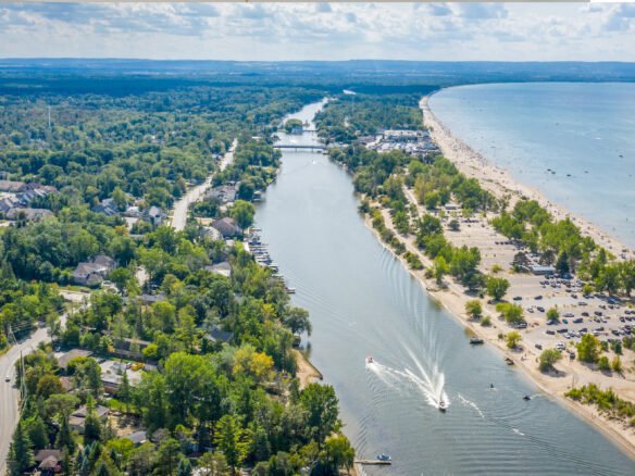 Aerial view of a coastal town with a river running through it, lined with trees and houses. A sandy beach with parked cars and recreational boats on the water is visible. On the horizon, a lush landscape stretches under a partly cloudy sky, hinting at new homes emerging in this picturesque setting.