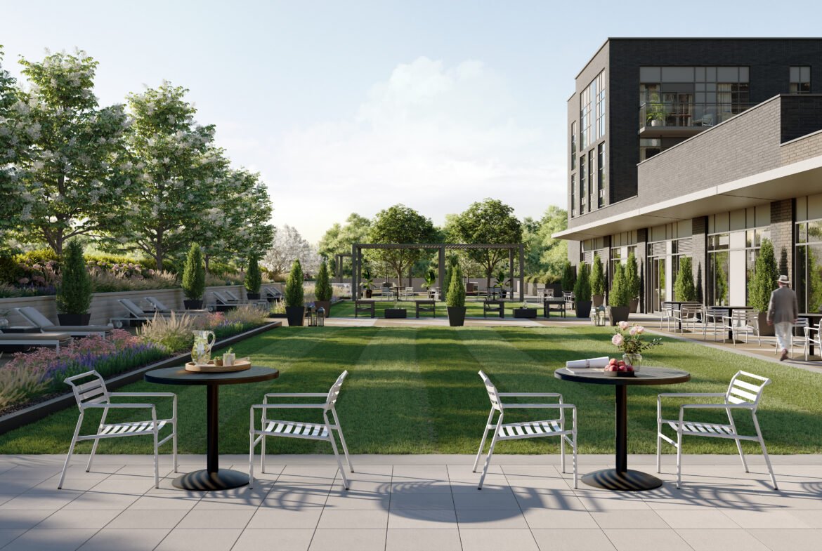 A modern outdoor patio with metal tables and chairs overlooks a landscaped garden filled with trees, plants, and a grassy area. On the right side, a contemporary building offers a glimpse of new condos in Brampton through its large windows. Sunlight filters through clouds, enhancing the serene atmosphere.