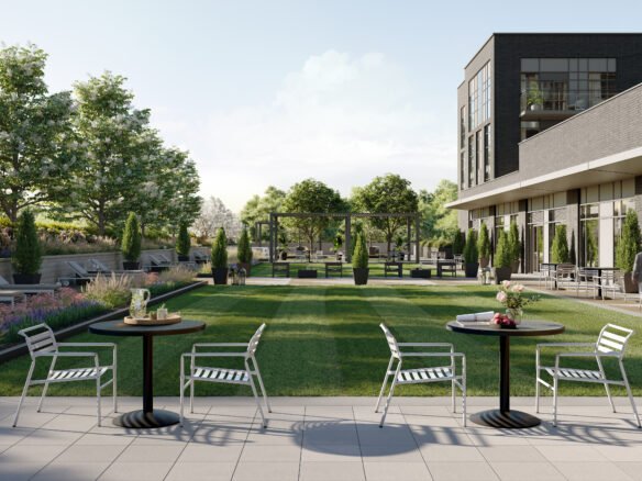 A modern outdoor patio with metal tables and chairs overlooks a landscaped garden filled with trees, plants, and a grassy area. On the right side, a contemporary building offers a glimpse of new condos in Brampton through its large windows. Sunlight filters through clouds, enhancing the serene atmosphere.