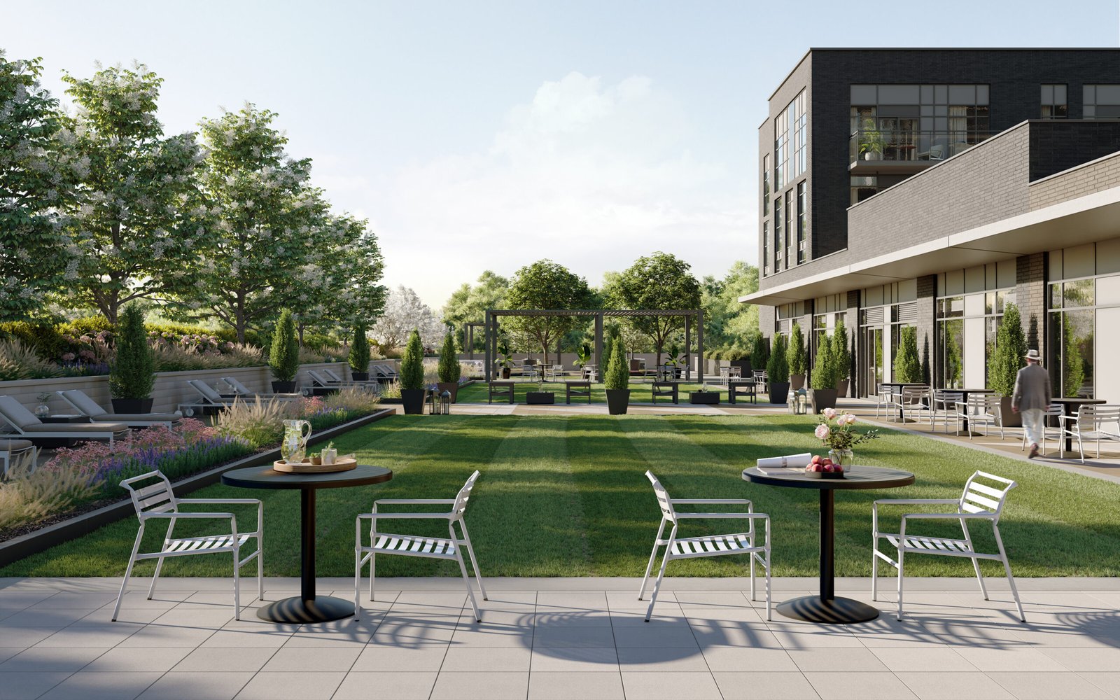 A modern outdoor patio with metal tables and chairs overlooks a landscaped garden filled with trees, plants, and a grassy area. On the right side, a contemporary building offers a glimpse of new condos in Brampton through its large windows. Sunlight filters through clouds, enhancing the serene atmosphere.