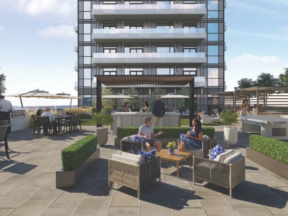 The rooftop terrace offers a lively space with people relaxing on patio furniture amidst lush plants. A modern building adds to the backdrop, while colorful umbrellas and tables dot the scene. The bright atmosphere showcases a clear sky, perfect for enjoying views of new condos in GTA.