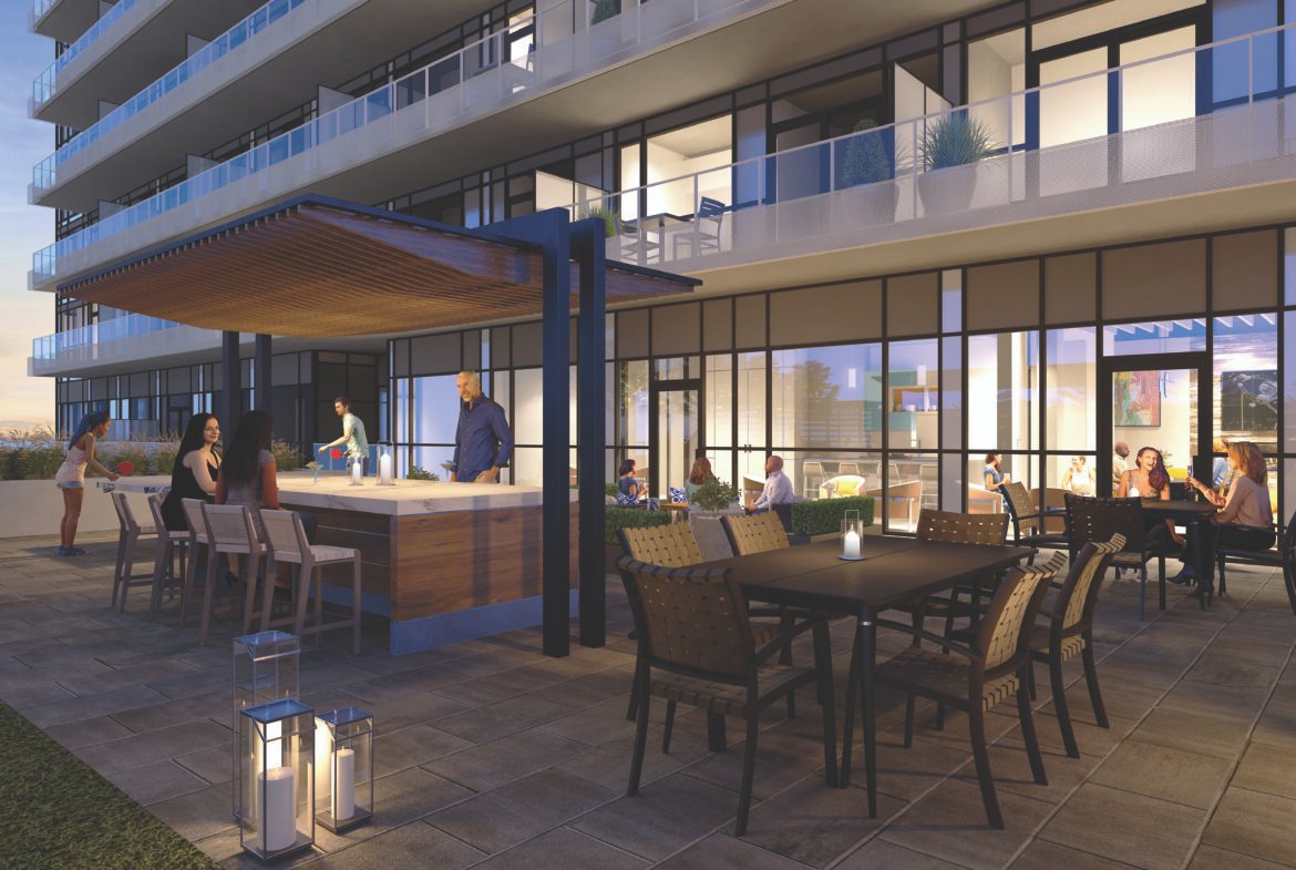 A modern outdoor patio at an apartment building with people socializing. The space features a covered bar area with stools, a dining table with chairs, and decorative lanterns. Large glass windows reveal a warmly lit interior, embodying the charm of new homes in Brampton.