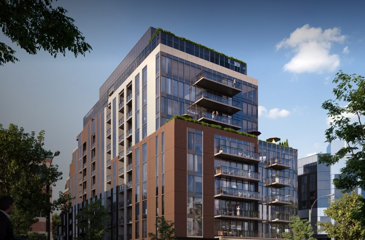 A modern multi-story building with large glass windows and balconies is surrounded by trees under a clear blue sky, creating a contemporary urban setting. Perfect for those interested in new condos in the GTA, this architecture promises stylish living amidst nature.