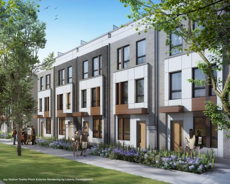 Joy Station Towns is a modern townhouse complex with three-story buildings featuring large windows and balconies. People stroll along the sidewalk bordered by trees and greenery on a sunny day.