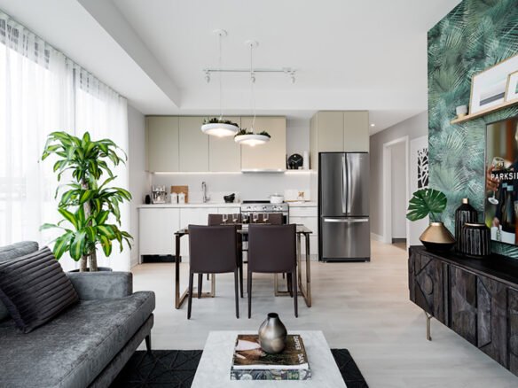 Modern living room with a gray couch, green plant, and coffee table. Open-concept kitchen in the background features a dining table, chairs, and stainless steel appliances. A TV on the right displays a scene. Decor includes leafy wallpaper and contemporary art, ideal for new homes in Toronto.