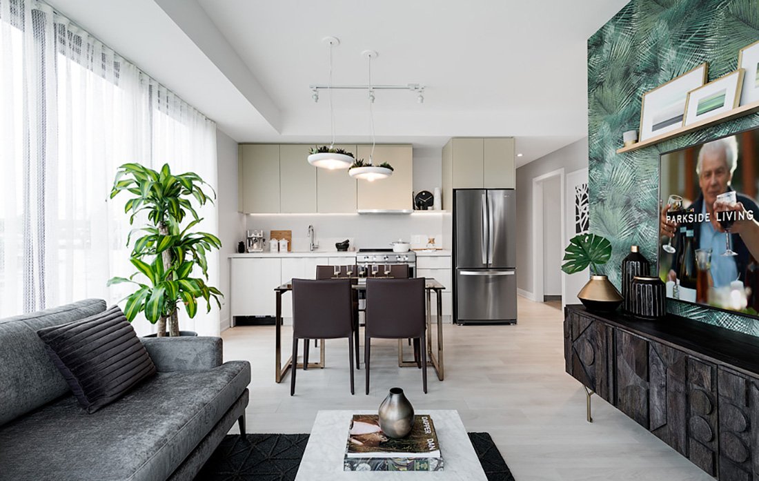 Modern living room with a gray couch, green plant, and coffee table. Open-concept kitchen in the background features a dining table, chairs, and stainless steel appliances. A TV on the right displays a scene. Decor includes leafy wallpaper and contemporary art, ideal for new homes in Toronto.