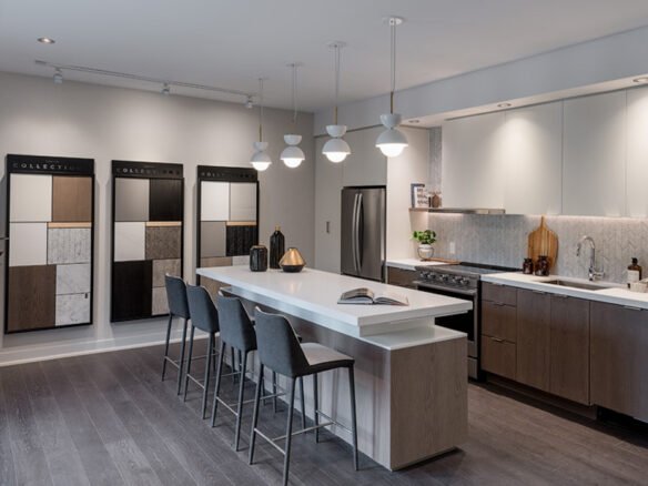Discover a modern kitchen showroom designed for pre-construction homes, featuring a central island, bar stools, and pendant lights. Sleek countertops and cabinetry complement the wall displays of materials and color samples. Neutral tones and contemporary elements create a sophisticated atmosphere.