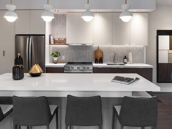 Discover the allure of a modern kitchen in new condos in Brampton, featuring a large white island, gray chairs, and stainless steel appliances. White cabinets pair with decorative vases and art pieces to complement the textured backsplash. Hanging lights elegantly illuminate the sleek space.