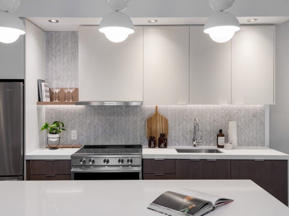 A modern kitchen with white cabinets, stainless steel appliances, and a patterned backsplash. Featuring a potted plant and cutting boards on the counter. Three hanging lights beautifully illuminate the space, perfect for new homes in Toronto. An open magazine rests on the island countertop.