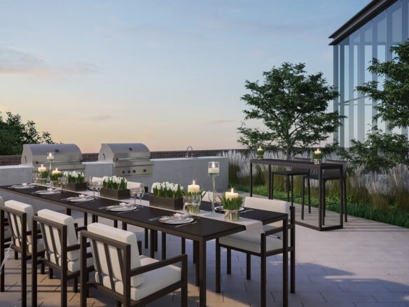 A modern outdoor patio in the new condos in Brampton features a long dining table with chairs, set for dinner with plates and candles. Two grills are on the side, and lush plants provide a scenic backdrop. The clear sky completes this serene evening setting.