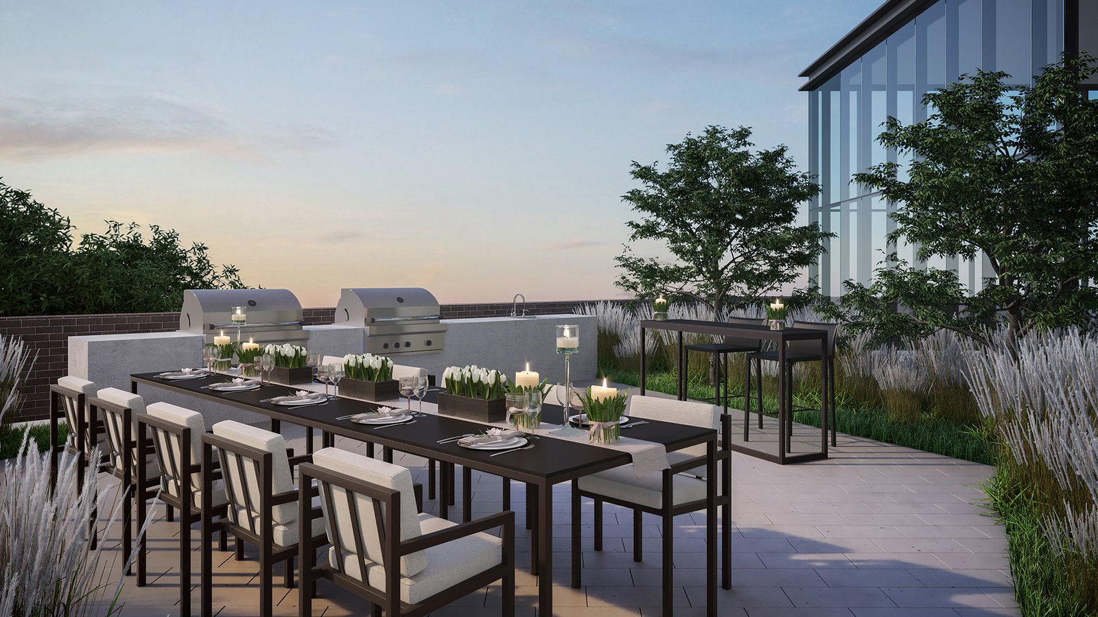 A modern outdoor patio in the new condos in Brampton features a long dining table with chairs, set for dinner with plates and candles. Two grills are on the side, and lush plants provide a scenic backdrop. The clear sky completes this serene evening setting.