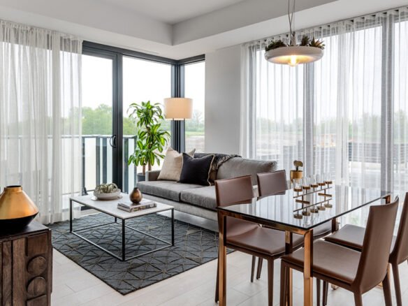Discover a modern living and dining room with large windows and sheer curtains, ideal for new homes. It features a grey sofa, coffee table, tall plant, dining table with four chairs, and a stylish light fixture. Light wood flooring and a decorative rug complete the space beautifully.