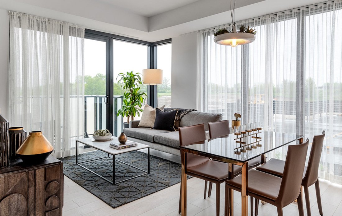 Discover a modern living and dining room with large windows and sheer curtains, ideal for new homes. It features a grey sofa, coffee table, tall plant, dining table with four chairs, and a stylish light fixture. Light wood flooring and a decorative rug complete the space beautifully.