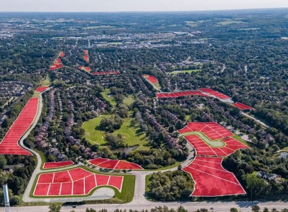 Aerial view of a suburban area with numerous houses surrounded by trees, featuring sections highlighted in red to indicate future development of pre-construction homes. Roads and lush greenery weave through this picturesque scene, promising vibrant new communities.