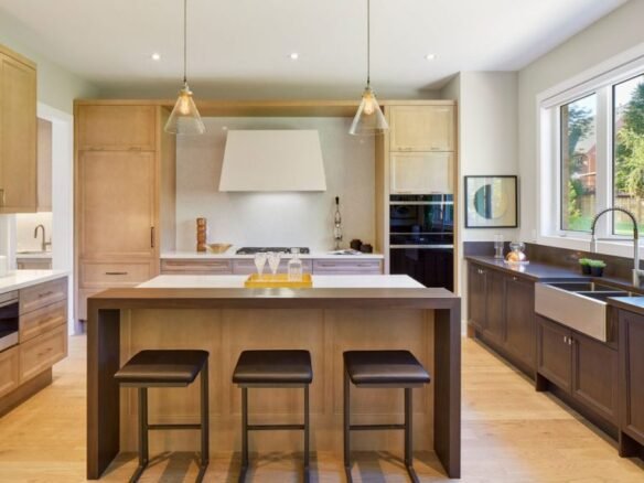 A modern kitchen in new homes features wooden cabinetry, a large island with three stools, pendant lights, and a built-in oven. A window above the double sink streams natural light into the room adorned in neutral tones.