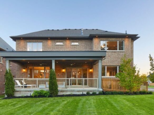 A charming two-story brick house with a wide wooden porch and pillars, surrounded by a well-maintained lawn and shrubs. Large windows grace both floors, basking in sunlight. It's the ideal inspiration for those considering new homes in GTA.