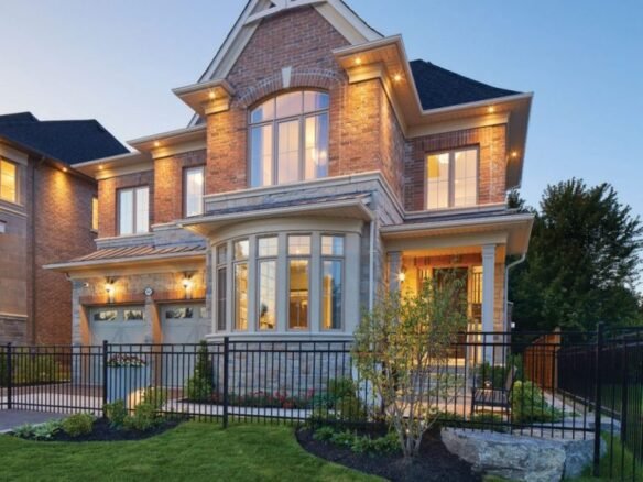 A large, modern brick house with a well-maintained lawn and a black iron fence epitomizes new homes in GTA. The residence features expansive windows, an illuminated porch, and two garage doors. As dusk settles in the clear sky, a warm glow elegantly bathes this stunning property.