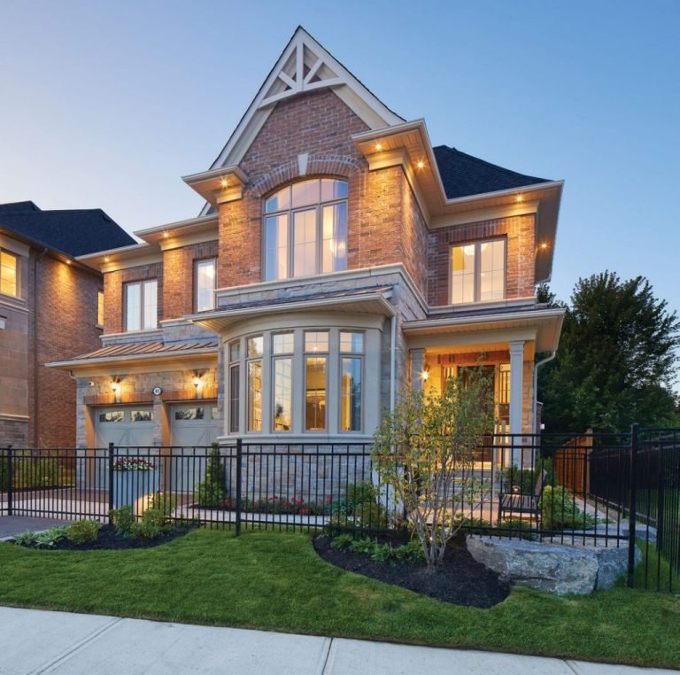 A large, modern brick house with a well-maintained lawn and a black iron fence epitomizes new homes in GTA. The residence features expansive windows, an illuminated porch, and two garage doors. As dusk settles in the clear sky, a warm glow elegantly bathes this stunning property.