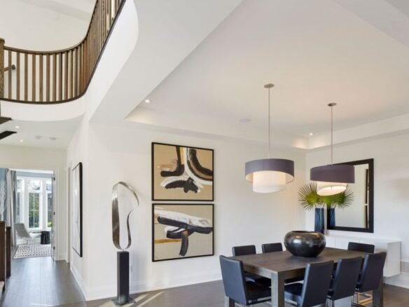 In a modern dining room typical of new homes in GTA, a dark wood table and six black chairs form the centerpiece. Two large abstract paintings adorn the white walls, complemented by pendant lights overhead. A unique sculpture graces the curved staircase railing as natural light floods the space.