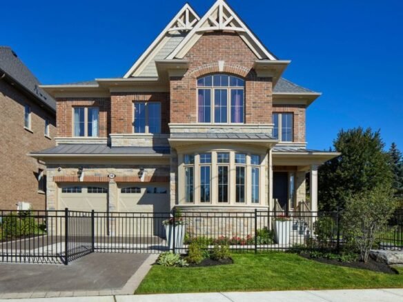 A two-story brick house with large windows and a gabled roof sits among new homes in the GTA. It features a double garage, manicured lawn, and a black metal fence. The sky is clear and blue, enhancing its modern charm.