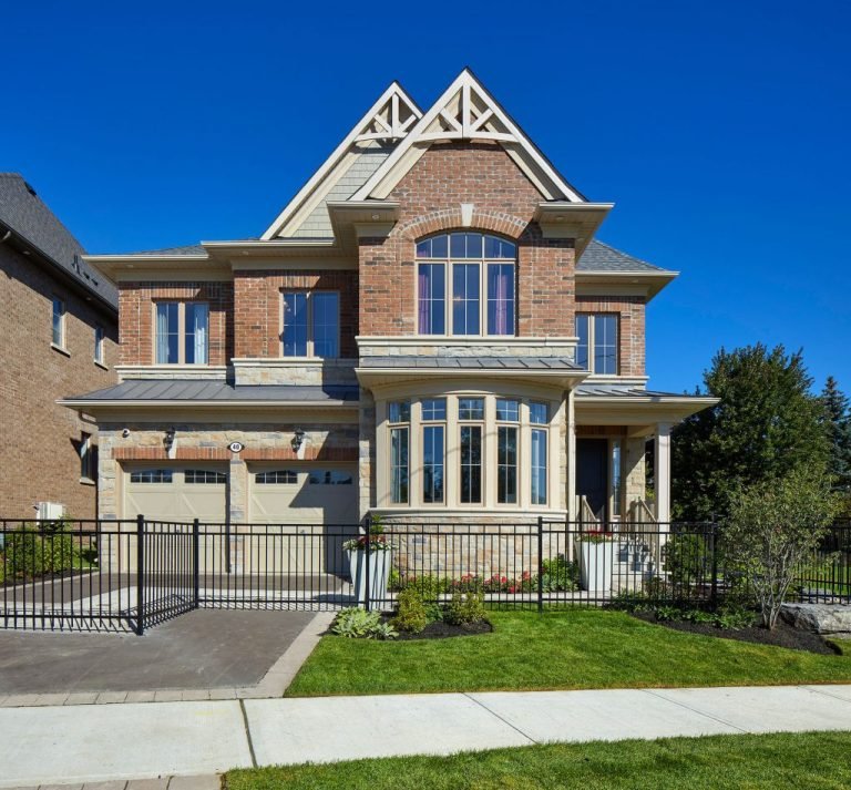 A two-story brick house with large windows and a gabled roof sits among new homes in the GTA. It features a double garage, manicured lawn, and a black metal fence. The sky is clear and blue, enhancing its modern charm.