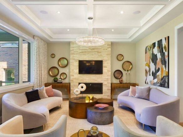 A modern living room in one of the new condos in GTA features two curved sofas facing each other, a central round coffee table, and a wall-mounted TV above a fireplace. With abstract wall art, decorative round mirrors, and a coffered ceiling, this space offers elegant comfort and style.