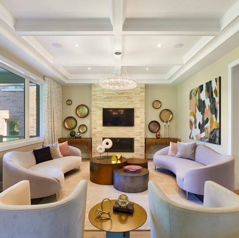 A modern living room in one of the new condos in GTA features two curved sofas facing each other, a central round coffee table, and a wall-mounted TV above a fireplace. With abstract wall art, decorative round mirrors, and a coffered ceiling, this space offers elegant comfort and style.