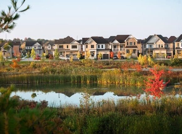 A row of modern suburban homes, part of the new homes in GTA, lines a street behind a pond reflecting the houses. Autumn foliage in reds, yellows, and greens surrounds the pond, while a clear blue sky stretches above.