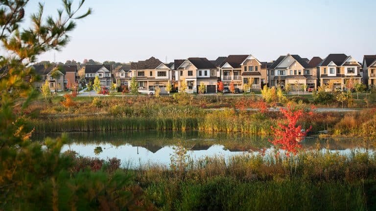 A row of modern suburban homes, part of the new homes in GTA, lines a street behind a pond reflecting the houses. Autumn foliage in reds, yellows, and greens surrounds the pond, while a clear blue sky stretches above.