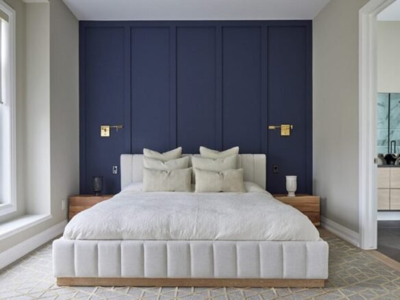 A modern bedroom in new condos features a large bed with white pillows and a cushioned headboard. The deep blue wall with gold accents enhances elegance, while natural light from the window highlights the room's luxurious feel. A glimpse of the bathroom completes this SG Homes masterpiece.