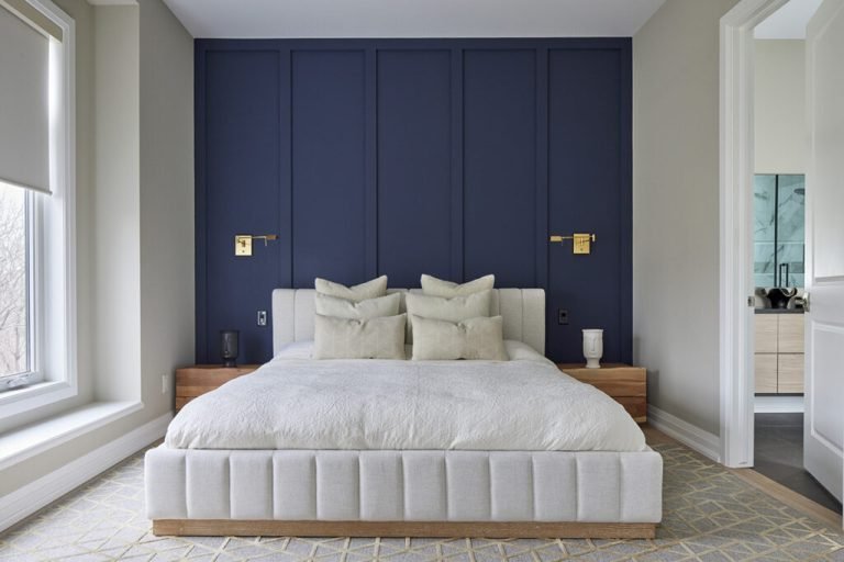 A modern bedroom in new condos features a large bed with white pillows and a cushioned headboard. The deep blue wall with gold accents enhances elegance, while natural light from the window highlights the room's luxurious feel. A glimpse of the bathroom completes this SG Homes masterpiece.