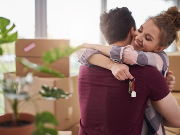 A woman hugs a man joyfully, holding a set of keys in her hand. Surrounded by cardboard boxes and the glow of a new beginning, they celebrate moving into their home. Despite the bustling housing market, they've found their place at a modest pace, with a potted plant gracing the foreground.