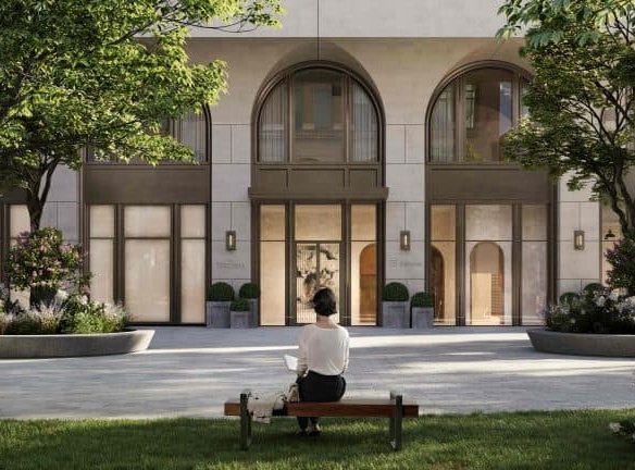 A person sits on a bench reading a book in front of the 101 Spadina Condos, a modern building with large arched windows and a central entrance. The surrounding area features trees and greenery, contributing to a serene, park-like setting. The weather appears clear and sunny.