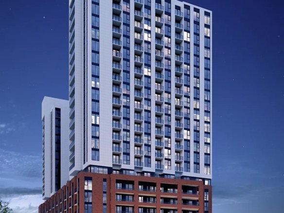 1989 Condominium A tall, modern apartment building from 1989 with a combination of red brick and white concrete facade, illuminated at night. Multiple windows and balconies are visible. The surrounding area has small trees and parked cars, with a twilight sky in the background.