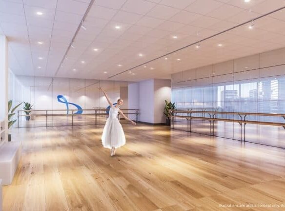 A dancer in a white ballet dress practices gracefully in a spacious, well-lit studio with wooden floors and mirrored walls. Large windows overlook a cityscape dotted with new condos. Potted plants and a blue ribbon decoration add charm to this urban oasis.