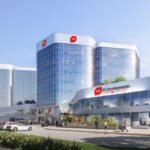 A modern office building with a reflective glass facade, labeled "NF Developments," stands amidst lush greenery and a bustling parking lot. It mirrors the ambitions of SG Homes in pre-construction projects. Above, a clear blue sky and distant airplane trail complete the scene.