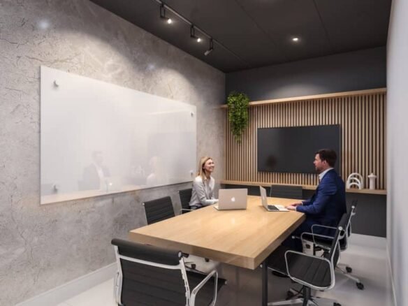 In a modern office meeting room with a long wooden table and four black chairs, two people sit with laptops facing each other. The room features a whiteboard on a textured wall and vertical plant decor. Ceiling lights illuminate the space, reminiscent of sleek designs found in new condos.