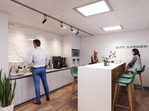 In the modern office kitchen of SG Homes, two women are seated at a tall table while a man in a white shirt and blue pants brews coffee. The space boasts white cabinets, wooden flooring, a plant, and the words "CITY GARDEN" elegantly displayed on the wall.