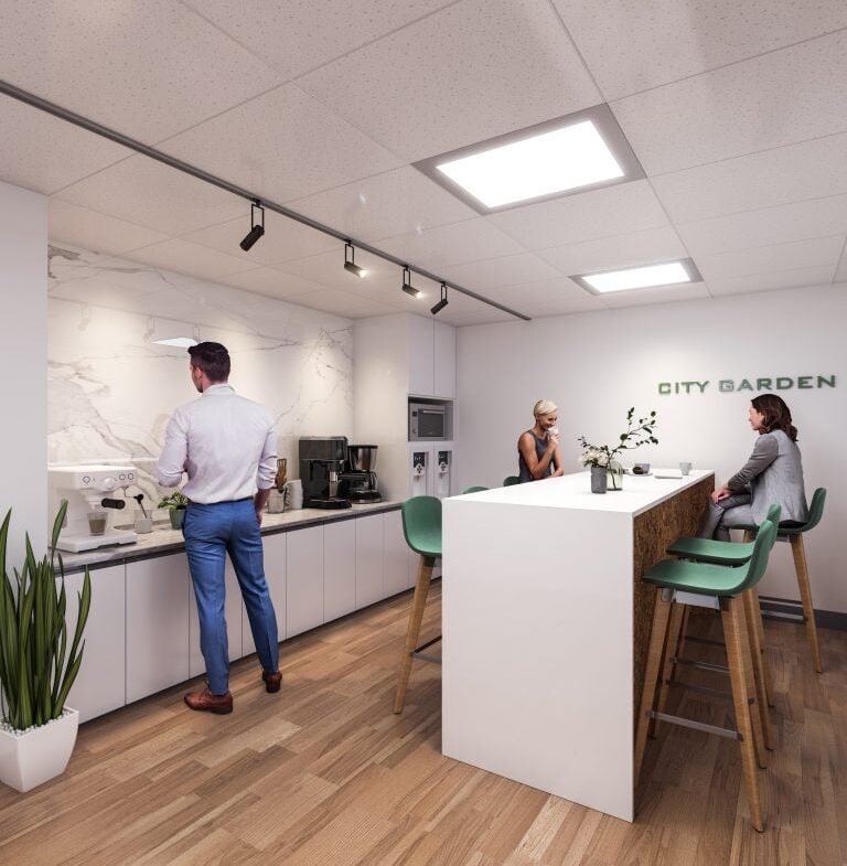 In the modern office kitchen of SG Homes, two women are seated at a tall table while a man in a white shirt and blue pants brews coffee. The space boasts white cabinets, wooden flooring, a plant, and the words "CITY GARDEN" elegantly displayed on the wall.