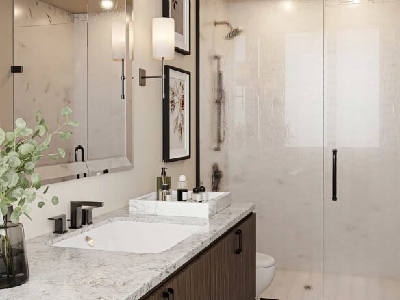 Modern bathroom in new GTA condos featuring a marble countertop with a sleek sink, a vase with greenery, and various toiletries. A large glass shower enclosure with a black handle is in the background, alongside framed artwork on the walls.