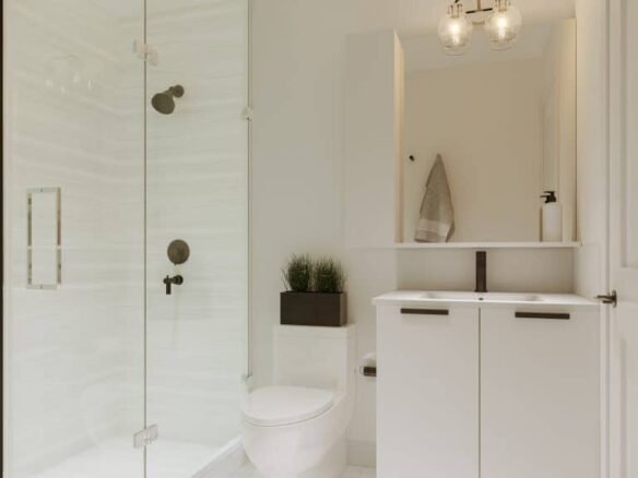 A modern bathroom in new GTA homes showcases a glass-enclosed shower with a black showerhead, a white vanity with black faucet, and a mirror above. A towel hangs neatly to the right, complemented by a potted plant on the shelf behind the toilet. Bright lighting enhances this stylish space.